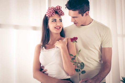 Beautiful happy young couple with red rose enjoying embracing near window
