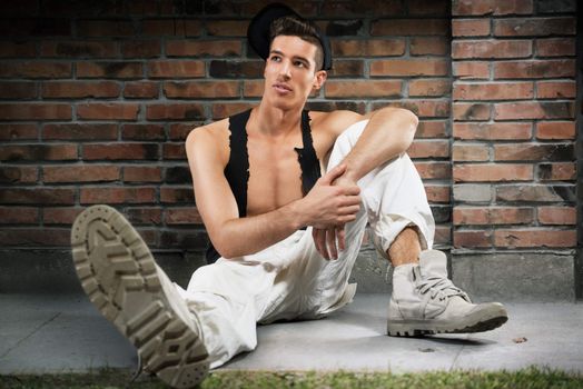 Portrait of young man in casual clothing and cap, sitting leaning against the wall and thinking. 
