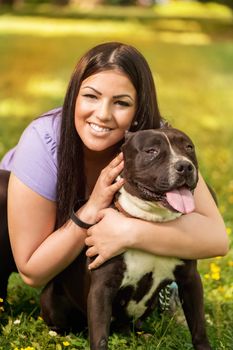 Portrait of a beautiful young woman hugging her cute stafford terrier. They sitting on grass in the park and enjoying. Looking at cemera.