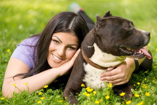 Beautiful young woman with her cute stafford terrier lying on grass in the park. Looking at camera. 