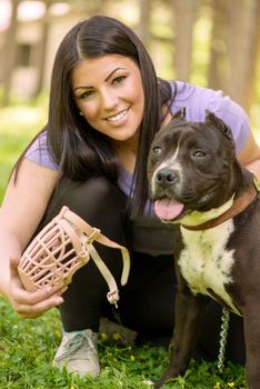 Beautiful young woman puts muzzle basket to her cute stafford terrier in the park .