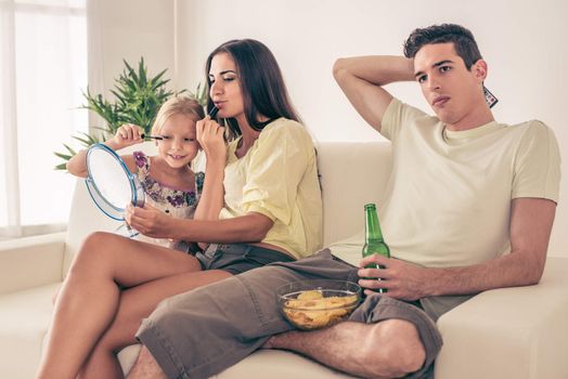 Young man sitting on sofa and watching football on tv. His wife and daughter having fun and make up.