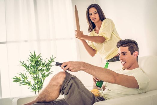 Young man sitting on sofa and watching tv. His wife is angry and standing with rolling pin.