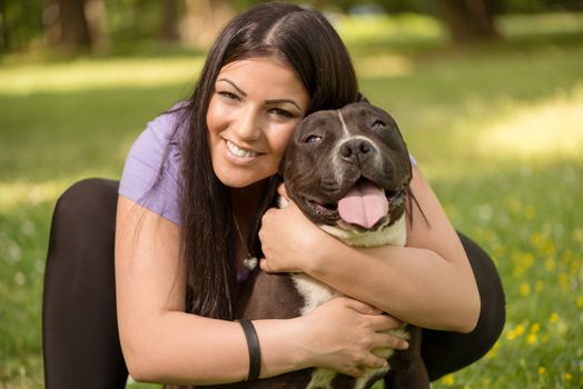 Portrait of a beautiful young woman hugging her cute stafford terrier in the park and enjoying. Looking at cemera.