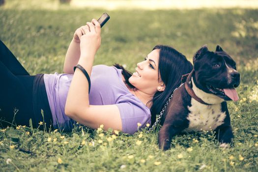 Beautiful young woman lying on the her dog in the park and typing a message on your mobile.