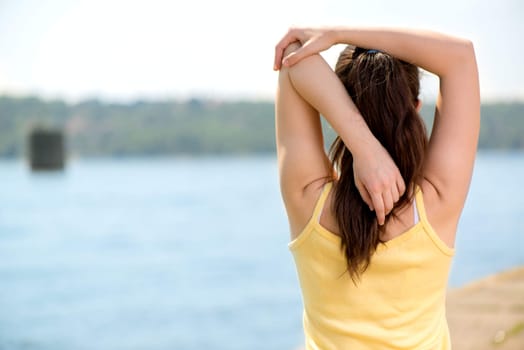 Beautiful young woman doing stretching exercise by the river. Rear view.
