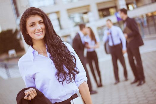 Portrait a young businesswoman standing in front of office building separated from the rest of the business team. Looking at the camera. 