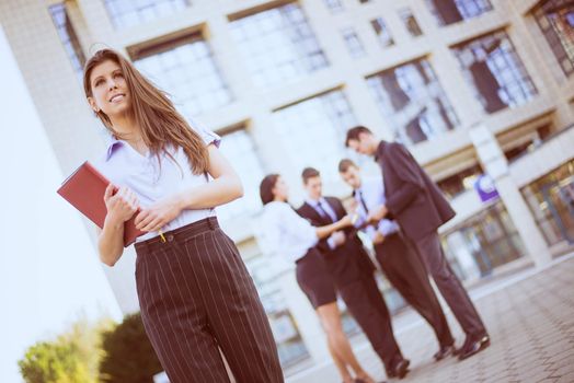 Young businesswoman with planner in her hands, standing in front of office building separated from the rest of the business team.
