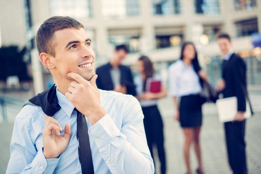 Portrait of a young thinking businessman standing in front of office building separated from the rest of the business team.