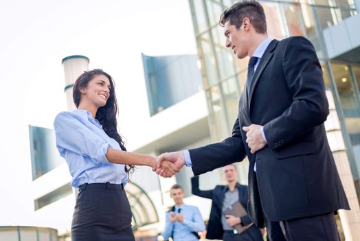 Young businesswoman and businessman shaking hands in front of the company while in the background standing theirs of the business team.