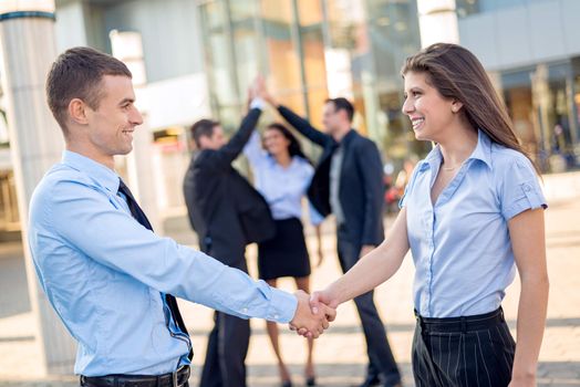 Young businesswoman and businessman shaking hands in front of the company while in the background standing theirs of the business team.