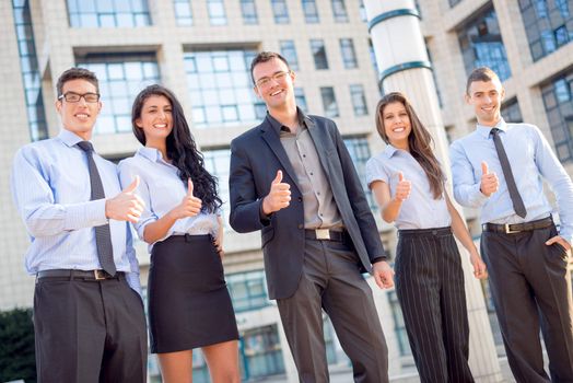 Young business team, elegantly dressed standing outside in front of office building with thumbs up. Looking at camera.