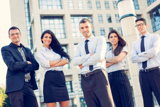 A small group of young business people standing in front of office building with arms crossed and with a smile on their faces looking at camera.