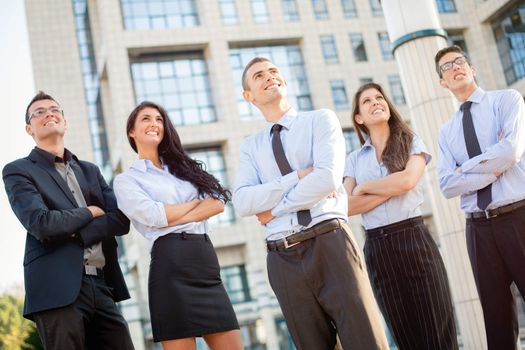 A small group of young business people standing in front of office building with arms crossed and with a smile on their faces looking in height.