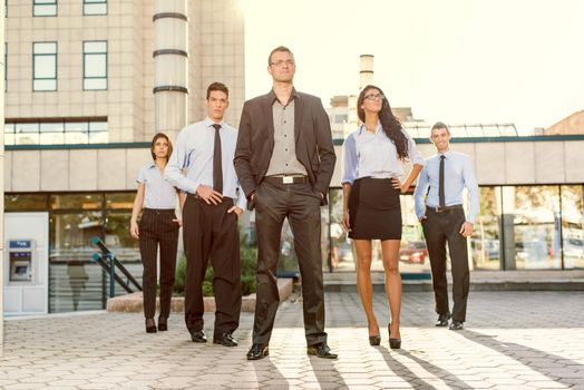 Young businessman, elegantly dressed with his hands in his pockets, standing proudly with his team of young businesswomen and businessmen in front of office building illuminated backlit sun.