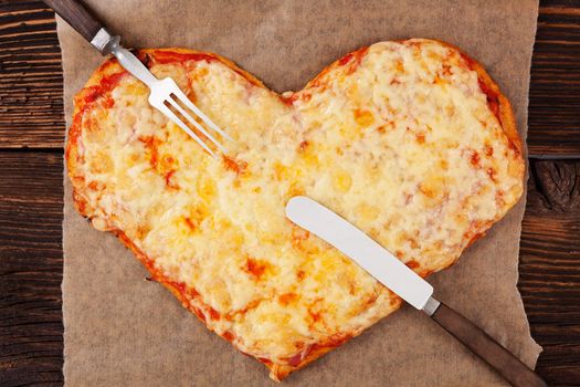 Margherita pizza in heart shape with cutlery on wooden table, top view. Culinary pizza eating. 