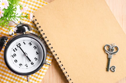 Focus at black alarm clock and brown diary with heart shape key on wooden background.