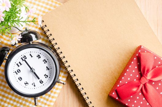 Alarm clock and brown diary with red gift box on wooden background.