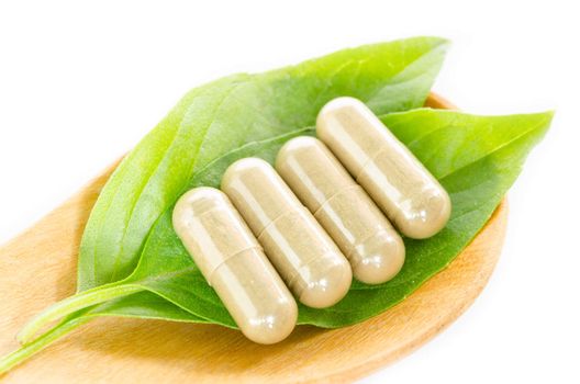 Yellow herb capsule with green leaf in wooden spoon on white background.