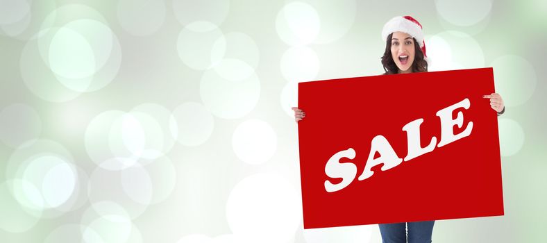 Pretty brunette in santa hat pointing white poster  against grey abstract light spot design