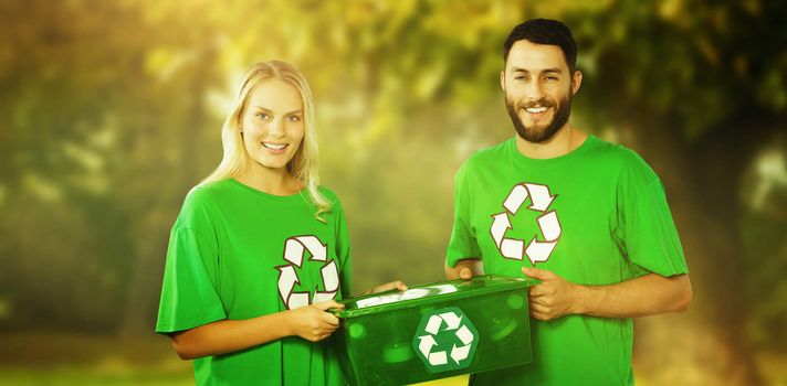 Portrait of smiling volunteers carrying recycling container  against trees and meadow in the park