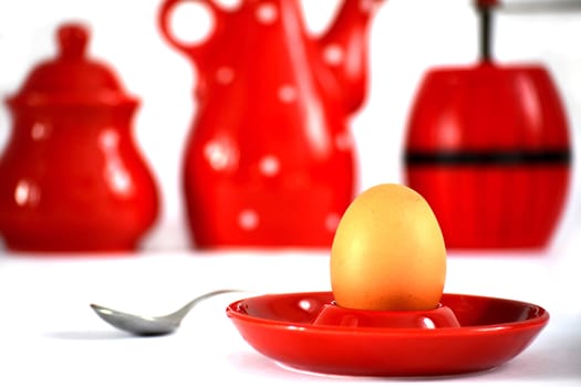 Egg on red holder near a spoon and cups and a pepper grinder in the background.