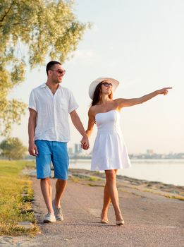 love, travel, tourism, gesture and people concept - smiling couple on summer vacation wearing sunglasses and pointing finger walking at seaside