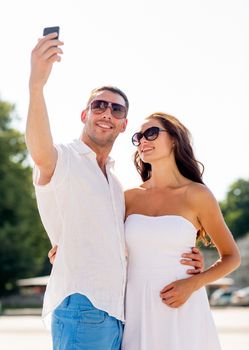 love, wedding, summer, dating and people concept - smiling couple wearing sunglasses hugging and making selfie with smartphone in park