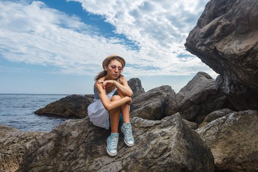 Beautiful young teen girl in dress and hat posing on the stones in the sea fashion shoot at shore