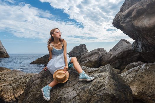 Beautiful young teen girl in dress and hat posing on the stones in the sea fashion shoot at shore