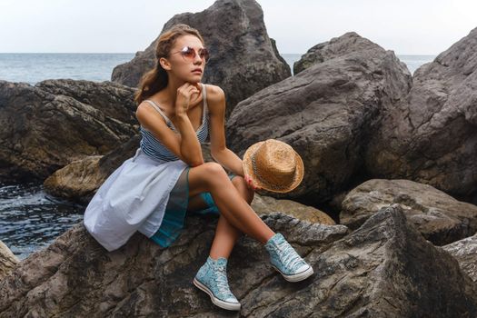 Beautiful young teen girl in dress and hat posing on the stones in the sea fashion shoot at shore