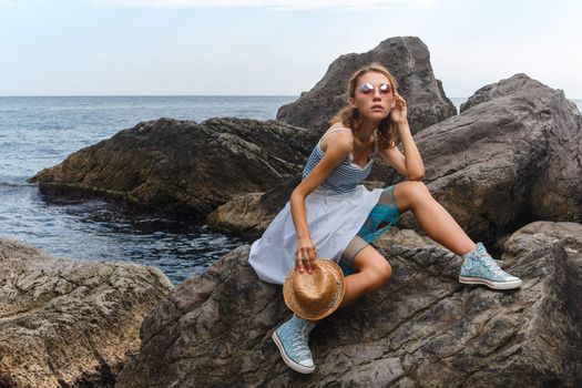 Beautiful young teen girl in dress and hat posing on the stones in the sea fashion shoot at shore