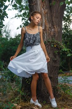 Fashion portrait shoot of a beautiful teen girl posing near tree during city fashion photoshoot with greenery at background