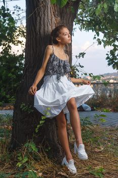 Fashion portrait shoot of a beautiful teen girl posing near tree during city fashion photoshoot with greenery at background