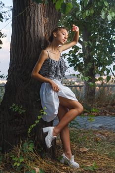 Fashion portrait shoot of a beautiful teen girl posing near tree during city fashion photoshoot with greenery at background
