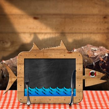 Blackboard in the shape of fish with blue waves and silver cutlery on a table with checkered tablecloth and fishing nets