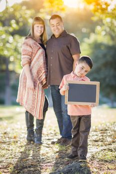 Young Mixed Race Couple Stands Behind Son with Blank Chalk Board Outdoors.
