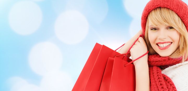 happiness, winter holidays, christmas and people concept - smiling young woman in hat and scarf with shopping bags over blue lights background