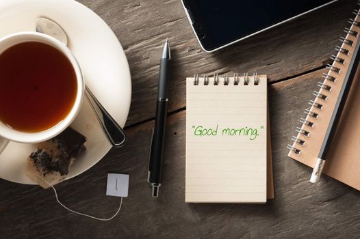 "Good morning" is written on small notepad with a cup of tea, pen, pencil and cellphne on rustic wood background in morning time with low key scene
