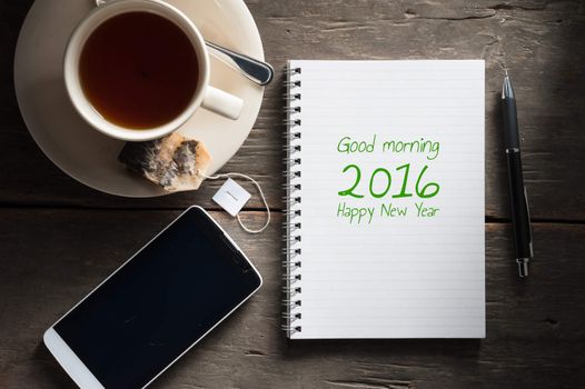 "Good morning 2016 Happy new year" is written on small notepad with a cup of tea, pen, pencil and cellphne on rustic wood background with low key scene