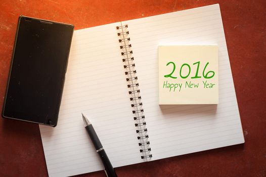 "2016 Happy new year" is written on opened notepad with pen, small sticky paper and black smart phone on rustic cement background with low key scene.