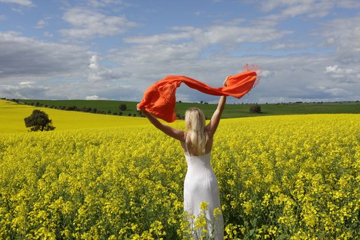 Happy woman wearing a long white dress and with scarf flailing in the air standing in a field of golden canola in rural Australia