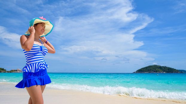 Beautiful woman in a blue striped swimsuit wear a hat standing and look at the sea and sky in summer on the beach of Koh Miang Island, Mu Ko Similan National Park, Phang Nga, Thailand, 16:9 widescreen