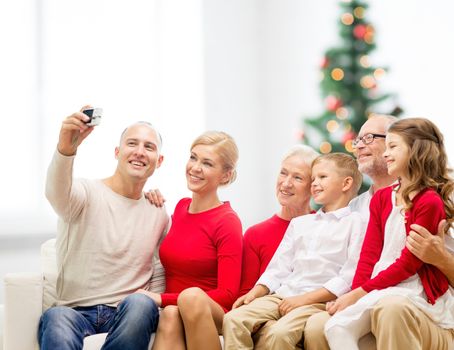 family, holidays, generation, home and people concept - smiling family with camera taking selfie and sitting on couch over living room with christmas tree background