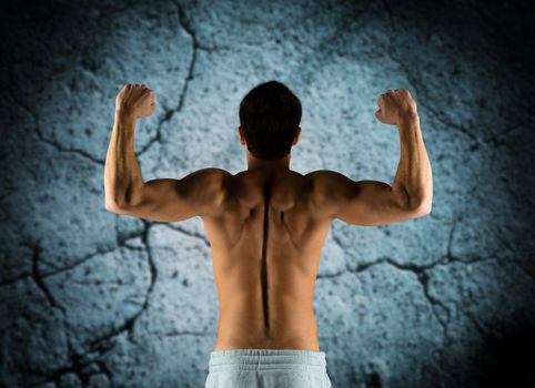 sport, bodybuilding, strength and people concept - young man showing biceps and muscles over concrete wall background