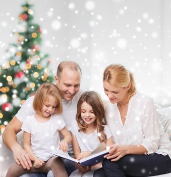 family, childhood, holidays and people - smiling mother, father and little girls reading book over living room and christmas tree background