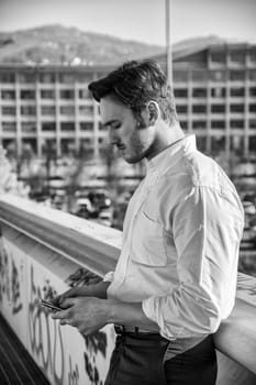 Handsome trendy man wearing white shirt standing and looking down at a cell phone that he is holding, outdoor in city setting