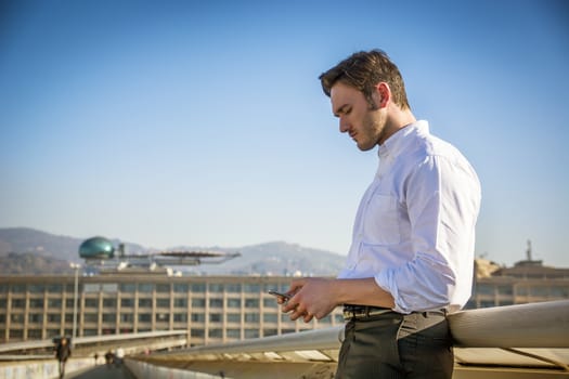 Handsome trendy man wearing white shirt standing and looking down at a cell phone that he is holding, outdoor in city setting