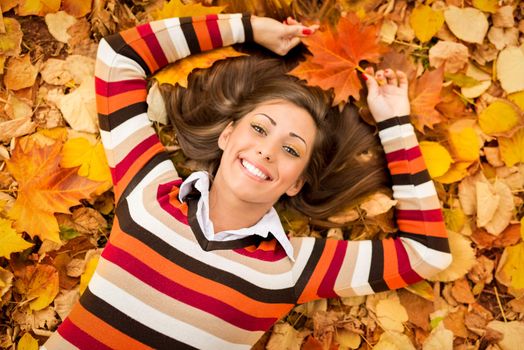 Beautiful young woman in sunny forest in autumn colors. She is lying on the fall leaf and enjoying. Looking at camera.