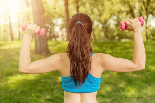 Beautiful girl doing exercise with dumbbell in the park. Rear view.